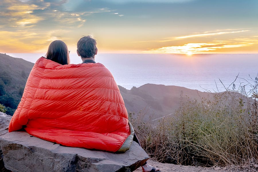 Couple with sleeping bag wrapped around them sitting on a rock watching the sun rise