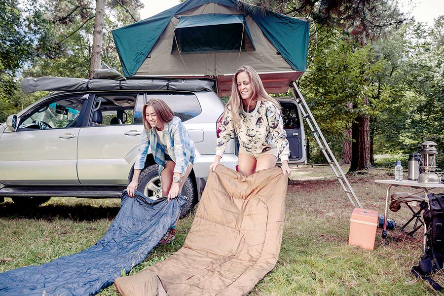 Two women with their sleeping bags in front of a vehicle with a roof top tent