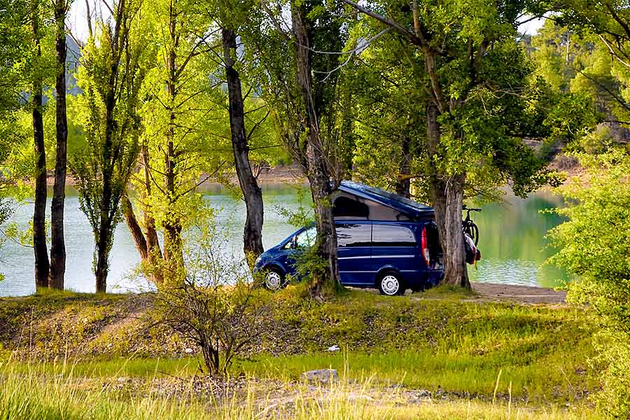 Blue pop top camper van parked under trees along side river