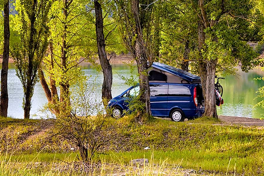 Blue camper van parked beside water in a wooded area