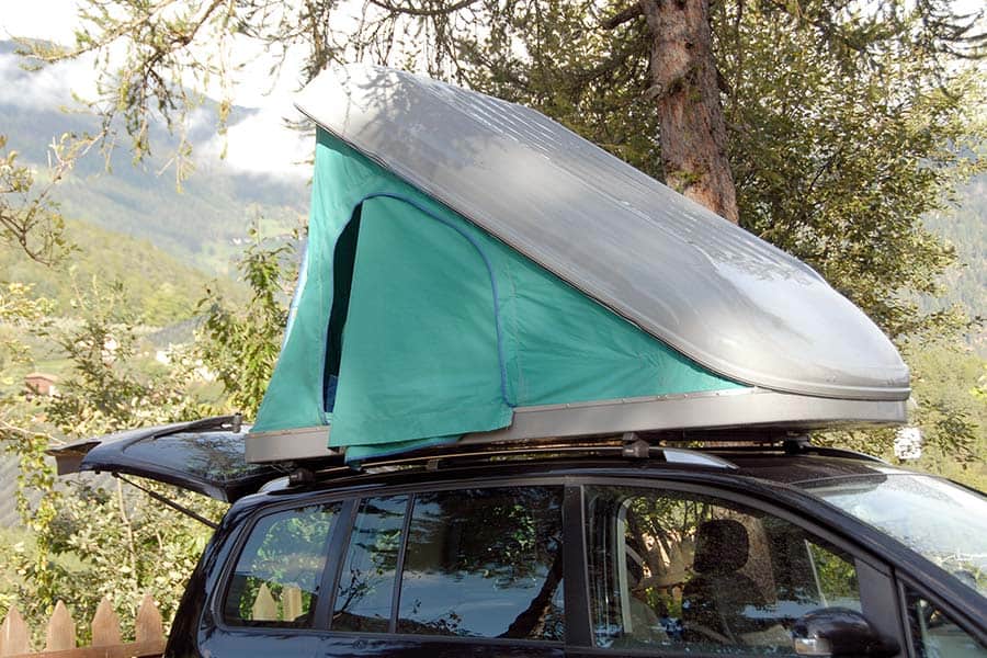 A green and silver rooftop tent on a dark colored SUV parked in a wooded area