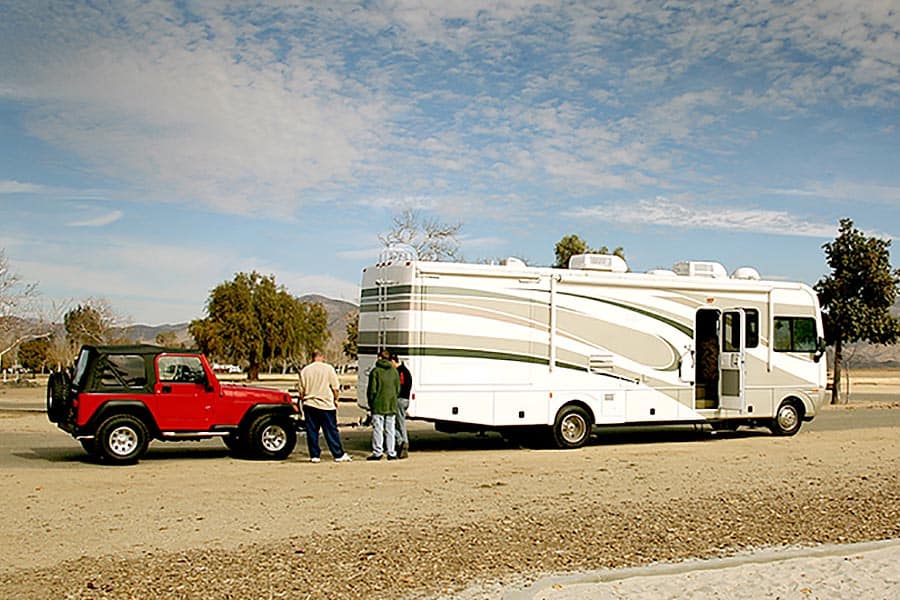 RV towing a red Jeep with three men looking at hitch